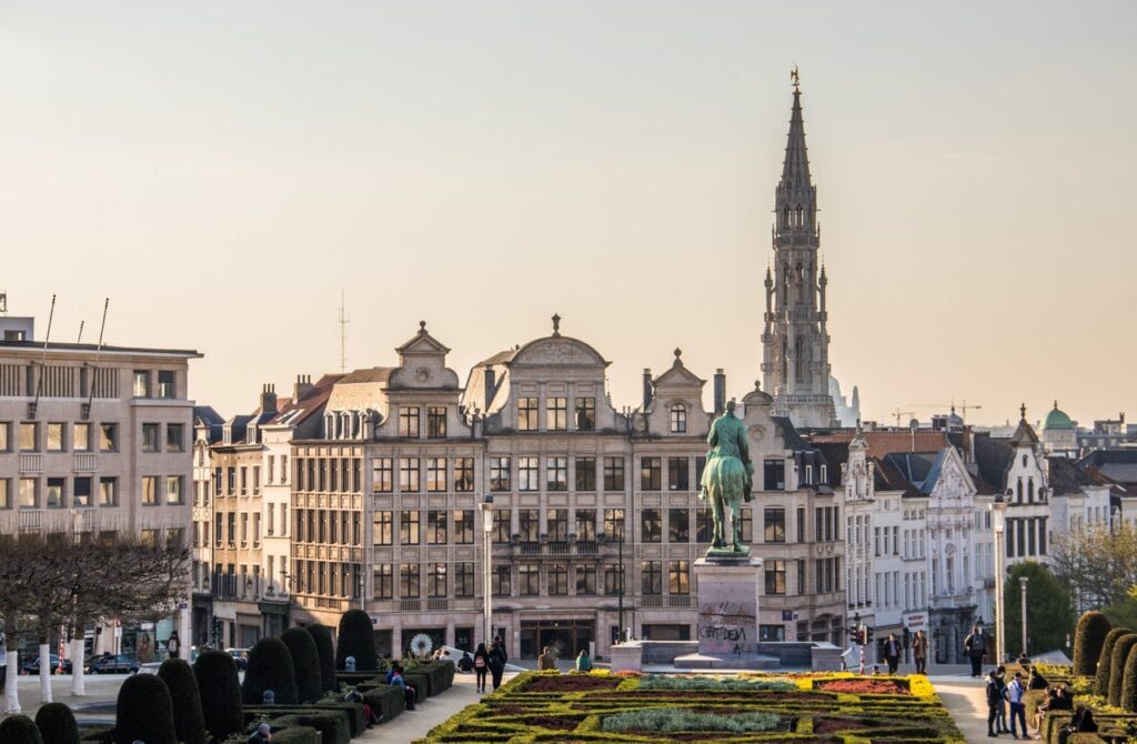 Vue panoramique de Bruxelles, en Belgique, avec le jardin du Mont des Arts au premier plan. La statue du roi Albert Ier à cheval se dresse en évidence. Des bâtiments historiques et la tour de l'hôtel de ville de Bruxelles s'élèvent à l'arrière-plan sous un ciel clair.