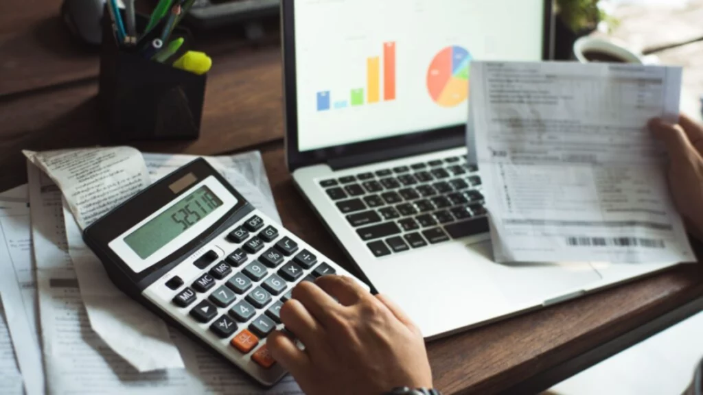 Personne calculant des finances avec une calculatrice et un ordinateur portable affichant des graphiques.