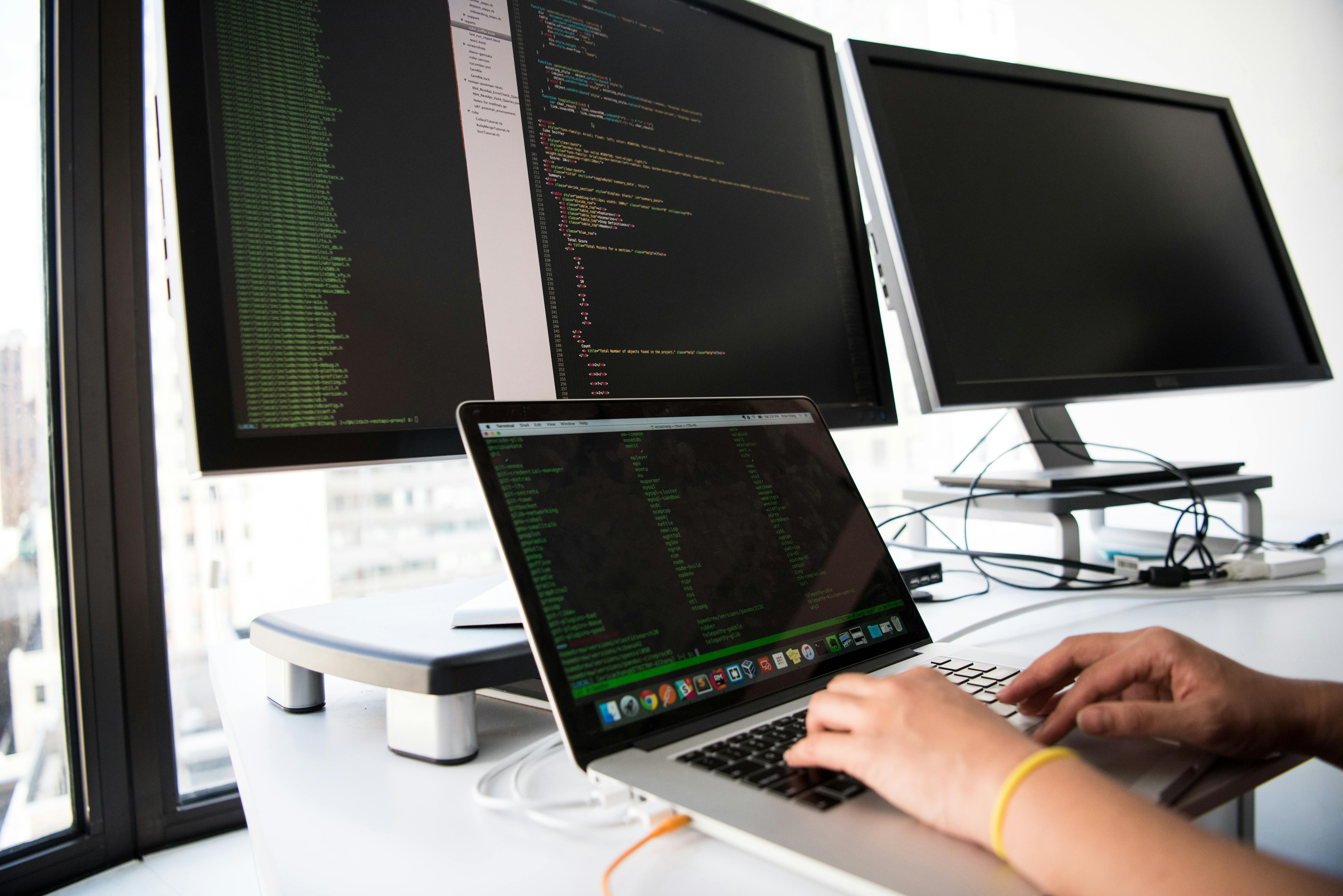 Développeur web travaillant sur un ordinateur portable avec deux grands écrans affichant du code.