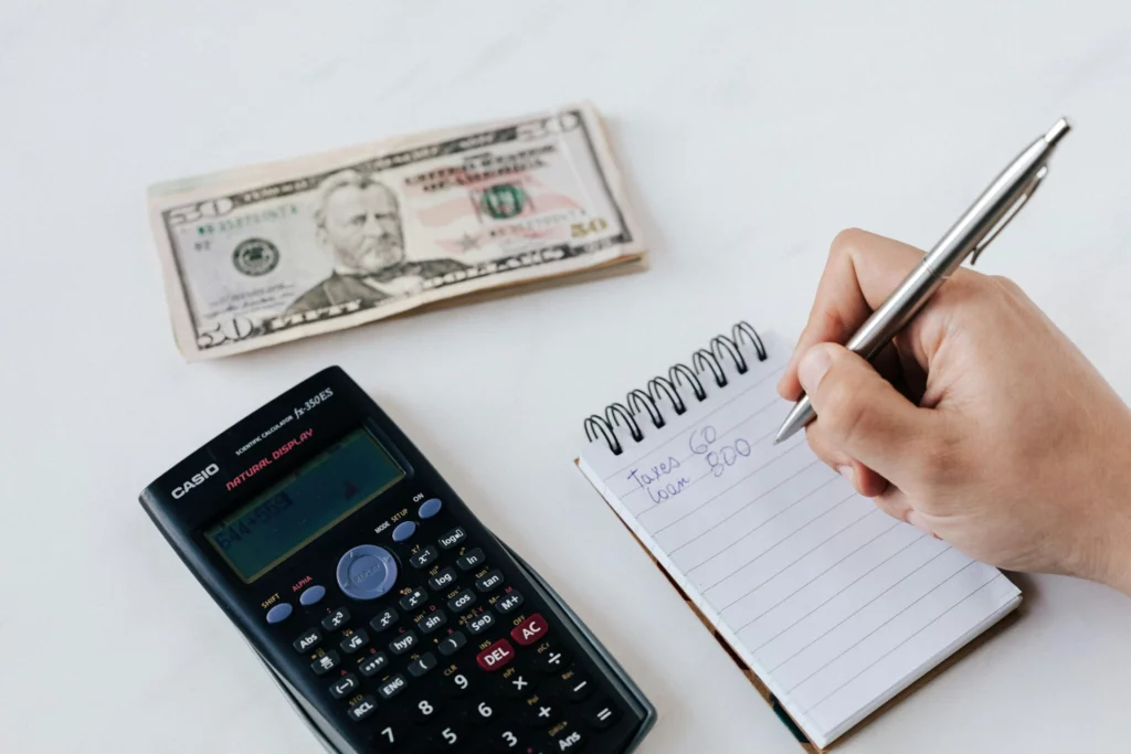 Main écrivant des calculs financiers sur un carnet à côté d'une calculatrice et de billets en dollars américains.