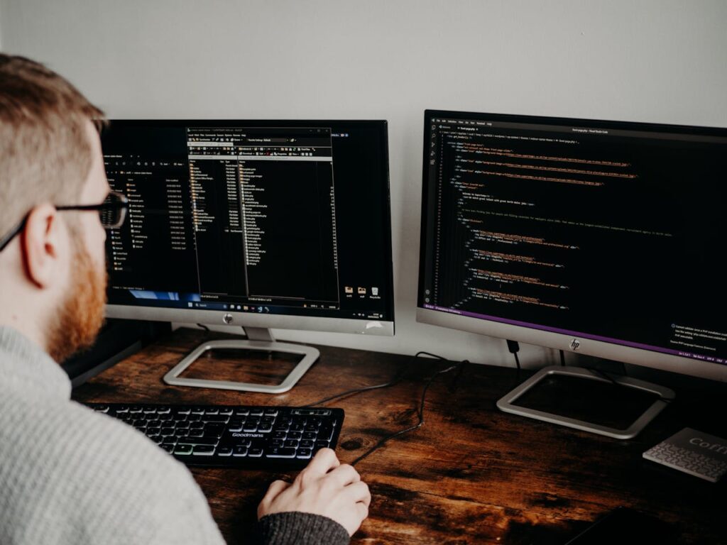 Un développeur web assis à son bureau, travaillant sur deux écrans affichant du code et l’arborescence de fichiers.