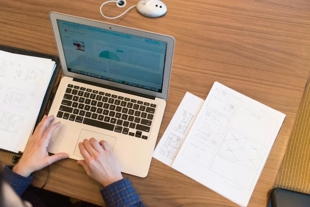 Conception d’interface web assistée par intelligence artificielle - iterates Utilisateur travaillant sur un ordinateur portable avec des wireframes papier sur un bureau.