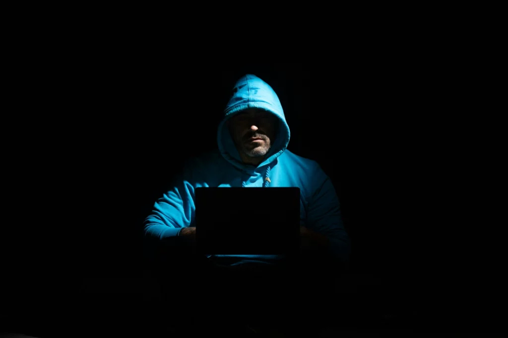 Pirate informatique masqué dans l’ombre - iterates Homme en sweat à capuche bleu utilisant un ordinateur portable dans l’obscurité.