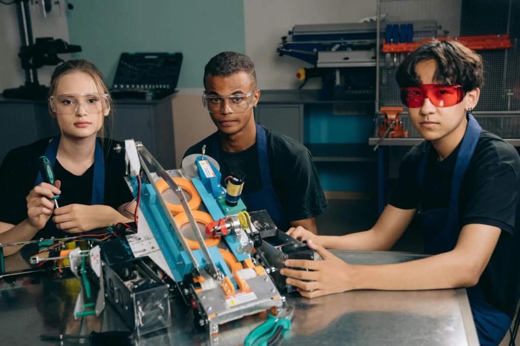 Trois jeunes portant des lunettes de protection travaillent sur un projet de robotique dans un atelier.