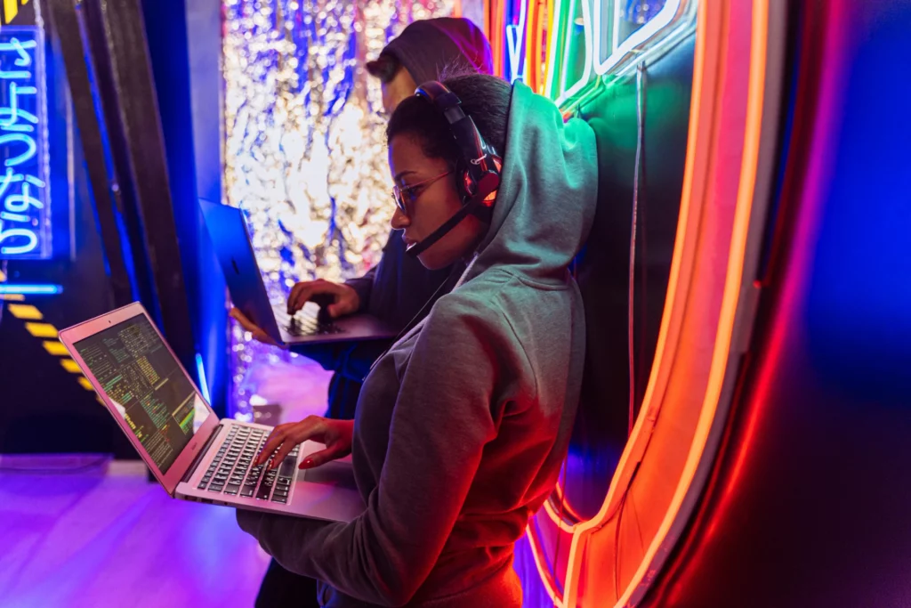 Hackers en action dans un environnement technologique coloré - iterates Femme en hoodie tapant sur un ordinateur portable avec des codes affichés à l’écran, accompagnée d’un collègue dans un environnement lumineux.