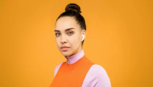 Une jeune femme portant des écouteurs sans fil, avec un pull orange et un fond jaune.