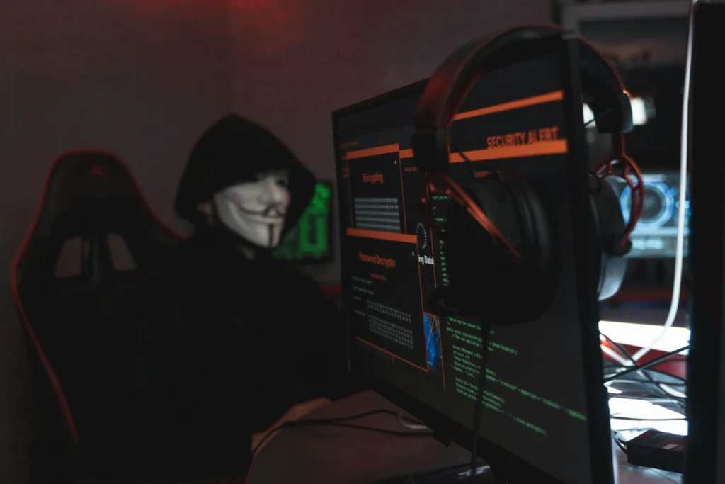 Hacker masqué devant un écran de sécurité - iterates Personne portant un masque de hacker devant un écran d’ordinateur affichant des alertes de sécurité.
