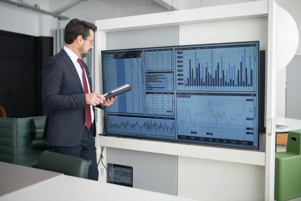 Analyse des données en temps réel - iterates Un homme d'affaires examine des données sur un écran à côté de lui tout en tenant une tablette, dans un bureau moderne.