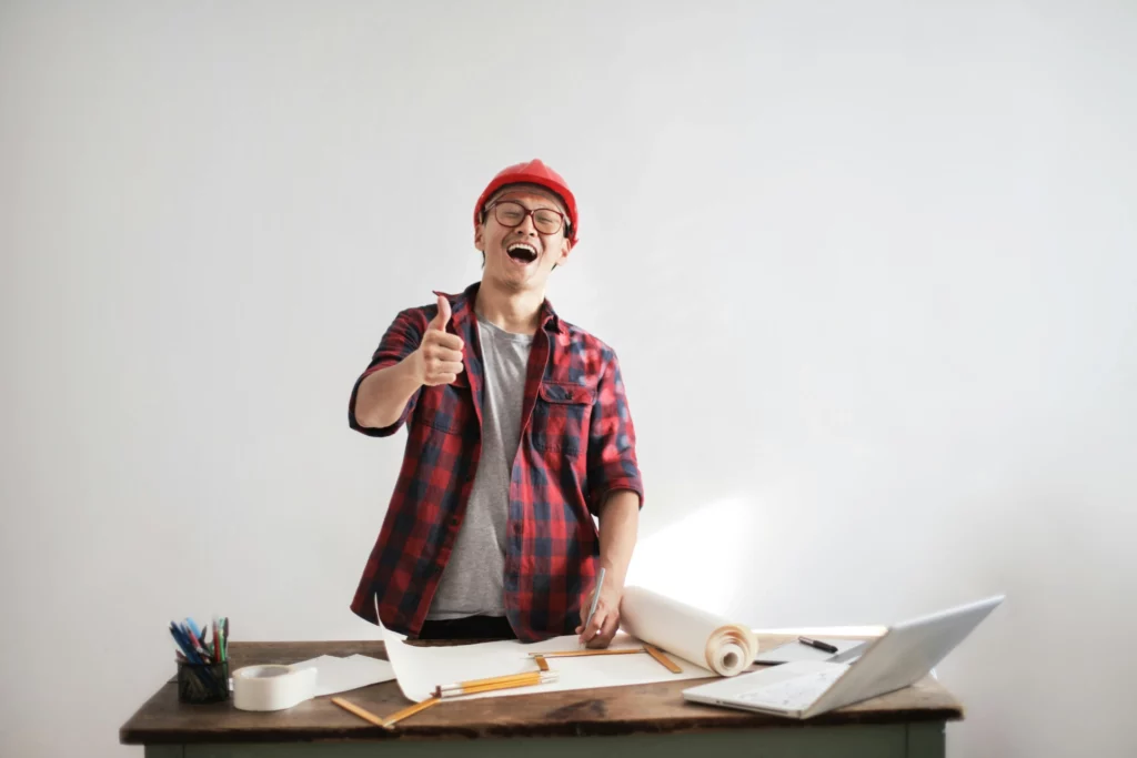 Succès d’un projet digital mené avec enthousiasme - iterates Homme souriant avec un casque de chantier levant le pouce devant un bureau de travail.