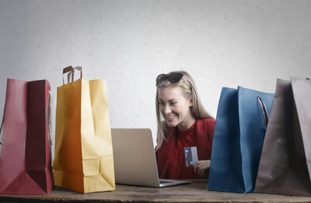 Achat en ligne réussi sur une boutique multivendeur - iterates Femme souriante faisant du shopping en ligne avec une carte bancaire, entourée de sacs d’achats.
