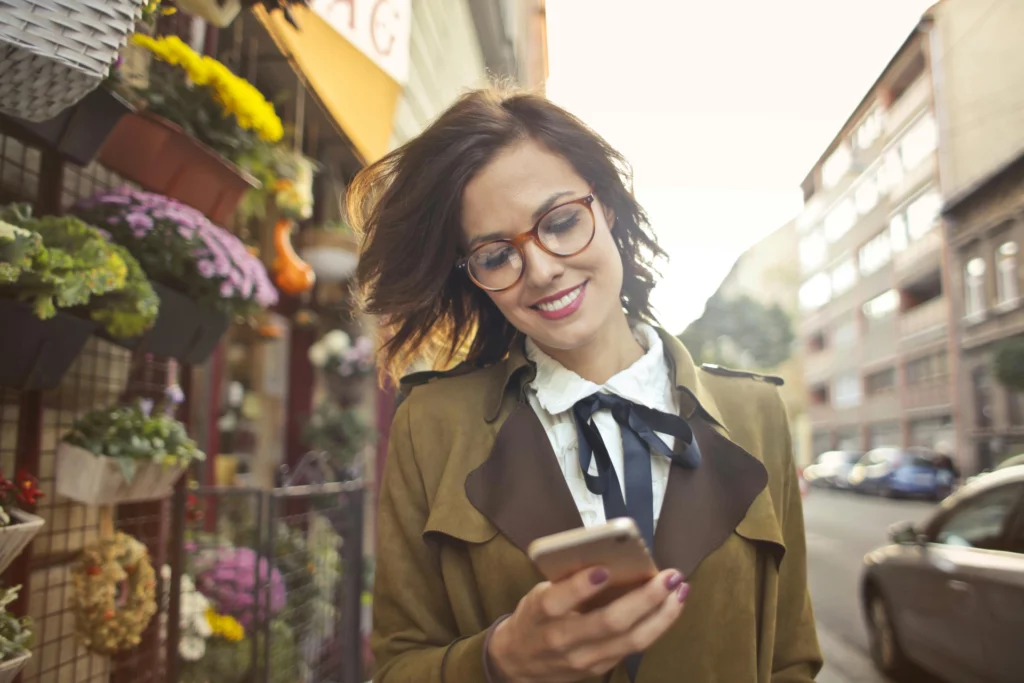Une jeune femme profite de sa promenade en ville pour consulter son téléphone, devant une boutique fleurie. - iterates Femme souriante portant des lunettes et consultant son smartphone en marchant dans une rue bordée de fleurs.