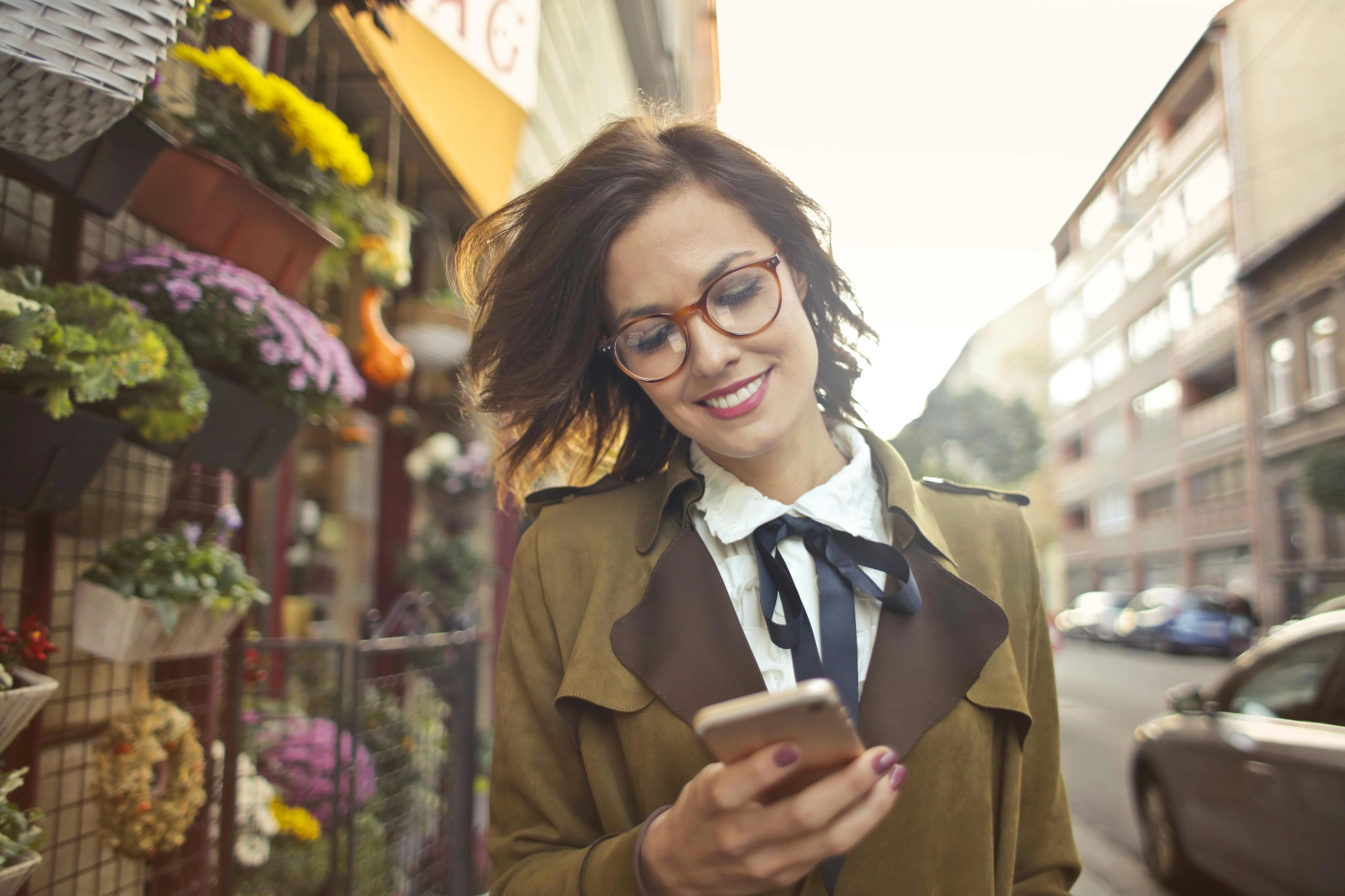 - iterates Smiling woman wearing glasses and consulting her smartphone as she walks along a flower-lined street.
