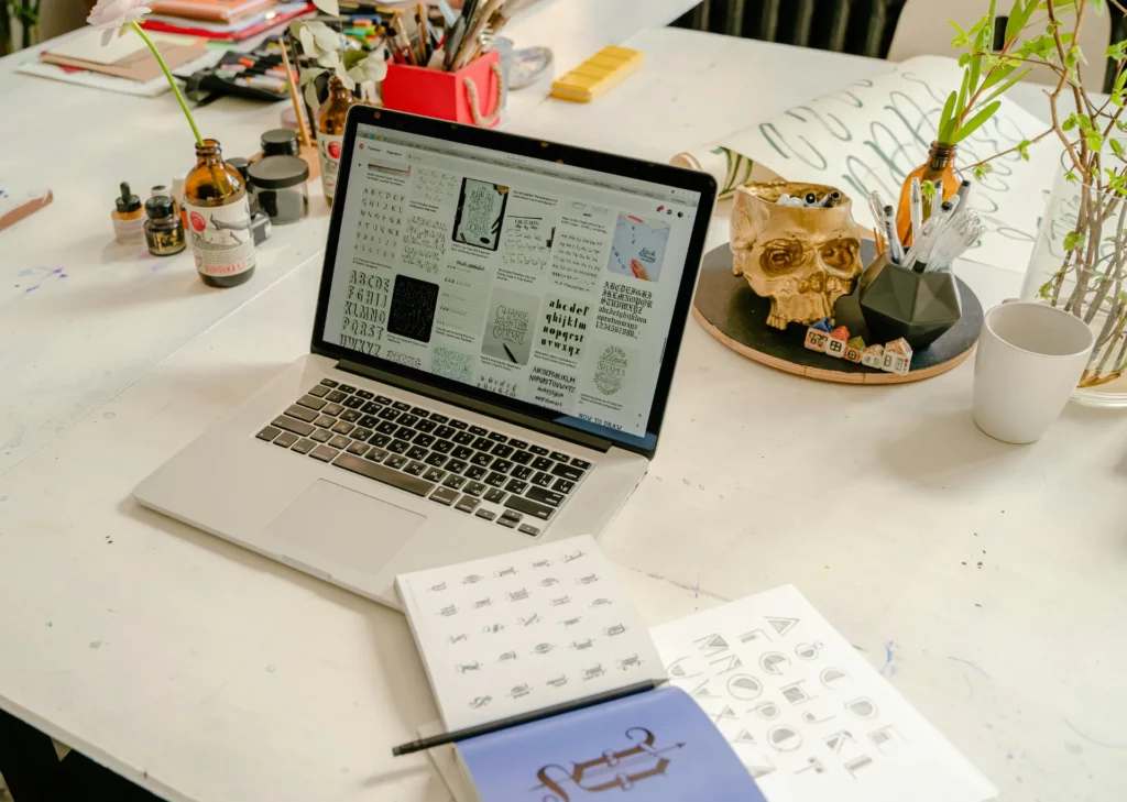 Ordinateur affichant des typographies sur un bureau de travail créatif avec carnets et outils de dessin.