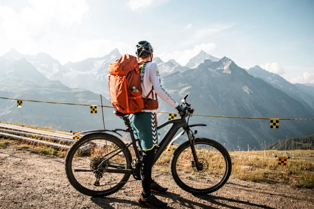 Vététiste face aux sommets alpins avec un sac à dos de randonnée - iterates Cycliste avec un sac à dos orange et un VTT contemplant un paysage de montagnes enneigées.