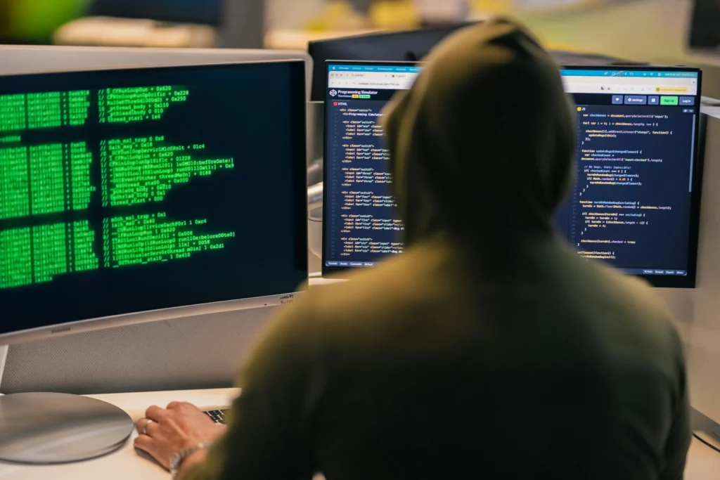 Cybercriminel en action sur un système informatique - iterates Personne en hoodie devant deux écrans affichant du code informatique et des données en vert.