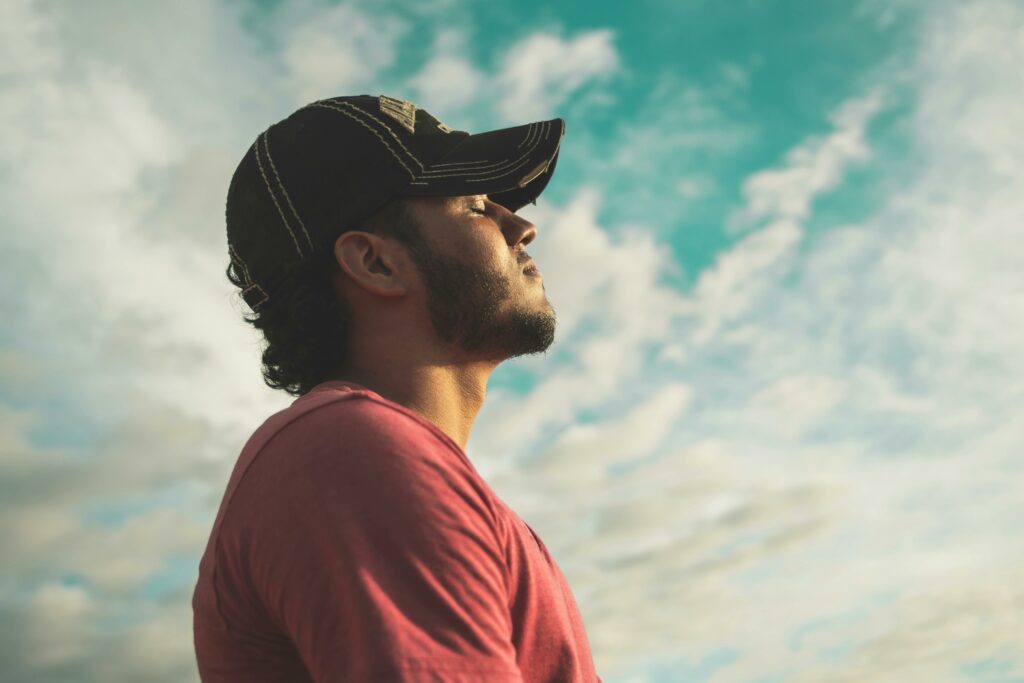 Pause mentale et sérénité après une crise - iterates Homme en casquette les yeux fermés, profitant d’un moment de calme sous un ciel bleu nuageux.
