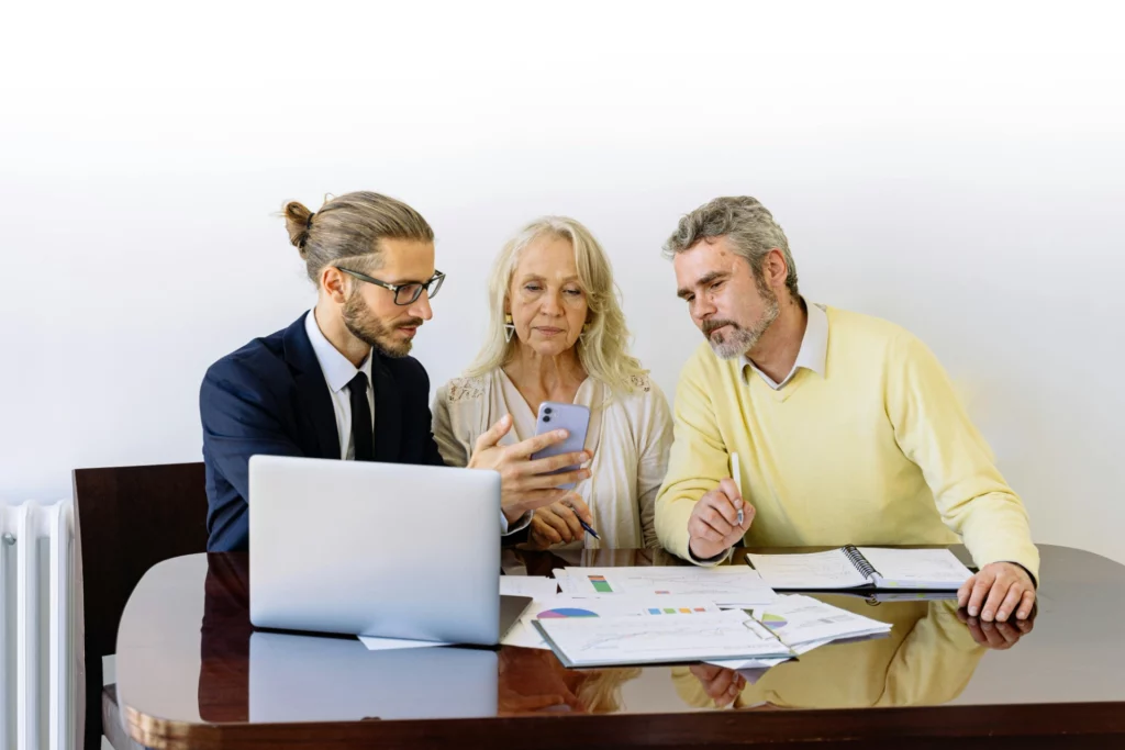Réunion professionnelle intergénérationnelle - iterates Un homme en costume, une femme âgée et un homme plus âgé, tous trois regardant un téléphone portable en discutant, autour d'une table avec des documents et un ordinateur portable.