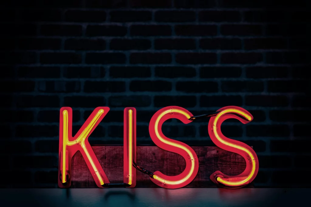 Signe au néon lumineux avec le mot "KISS" en rouge et jaune sur fond sombre de briques.