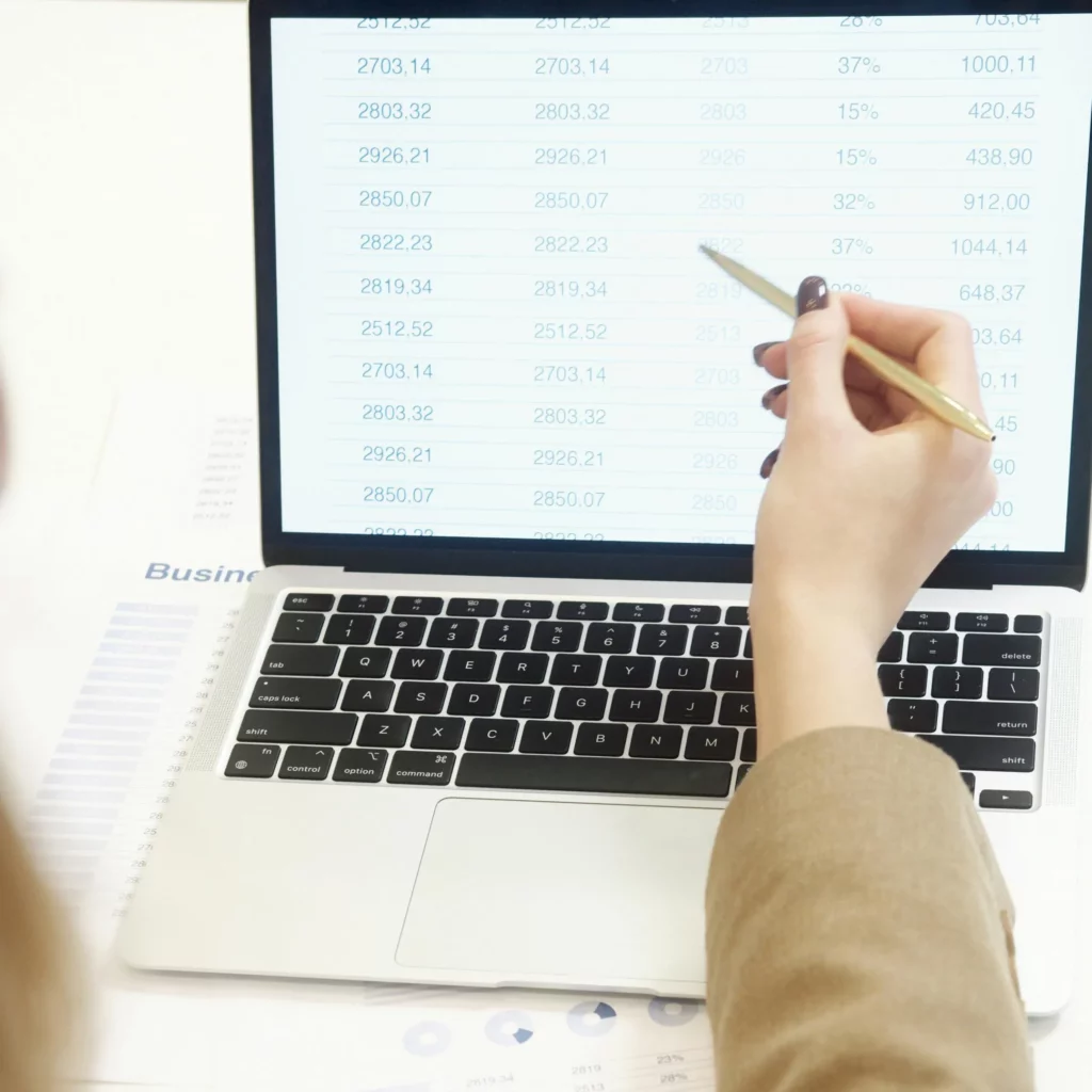 Close-up of a hand holding a pencil pointing at a spreadsheet with numbered rows on a laptop screen overlaid with documents.