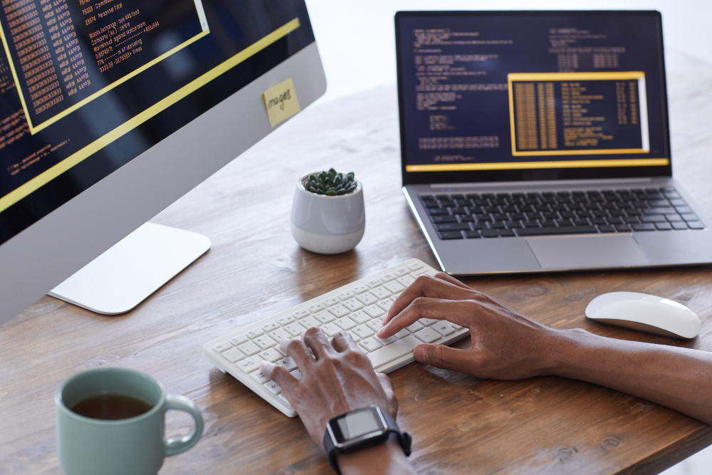 Hands typing on a white keyboard at a wooden desk with a desktop monitor and a laptop displaying code, plus a mug, a small plant, and a smartwatch nearby.