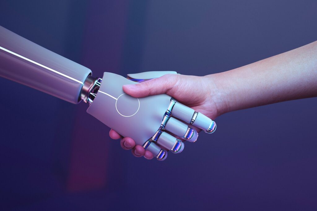 A human hand and a silver robotic hand clasp hands in a handshake against a dark blue gradient, symbolising collaboration.