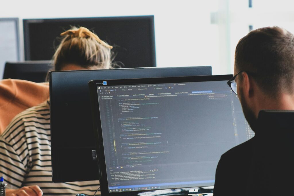 Two people coding at computer workstations in an office, concentrating on the code on their screens, with laptops nearby as part of a collaborative setup