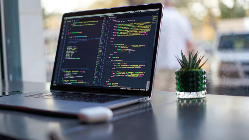 A laptop with colourful code on the screen is sitting on a desk next to a small potted plant.
