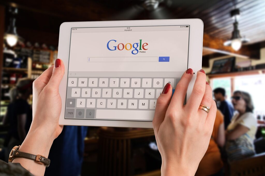 A person holding a tablet displaying the Google search page in a café setting, with a search bar visible on the screen and a keyboard shown at the bottom of the screen